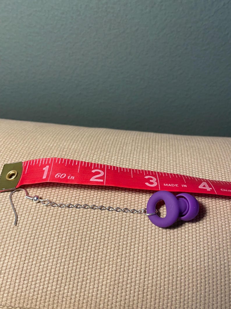 May include: A purple earplug earring featuring a silver-toned metal chain and a standard hook finding. The earring is positioned alongside a red measuring tape, which indicates the total length of the piece is approximately 3.5 inches long.