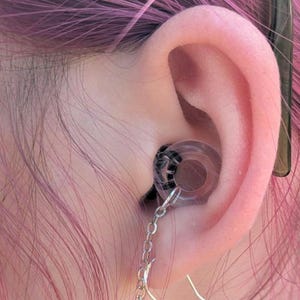 May include: A clear circular ear plug with a black spiral pattern inside, featuring a silver-toned metal chain attached to a hook earring component. The jewelry is inserted into the earlobe, with the chain dangling downward from the plug.