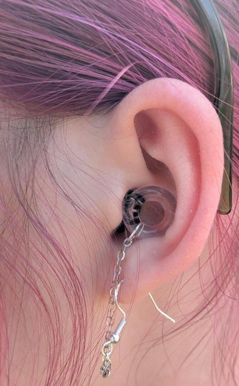 May include: A clear circular ear plug with a black spiral pattern inside, featuring a silver-toned metal chain attached to a hook earring component. The jewelry is inserted into the earlobe, with the chain dangling downward from the plug.
