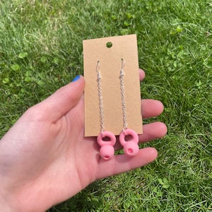 May include: A pair of dangle earrings featuring silver-toned metal chains attached to pink kettlebell-shaped charms. The earrings are displayed on a rectangular brown cardboard jewelry card with a small circular hole at the top for hanging.