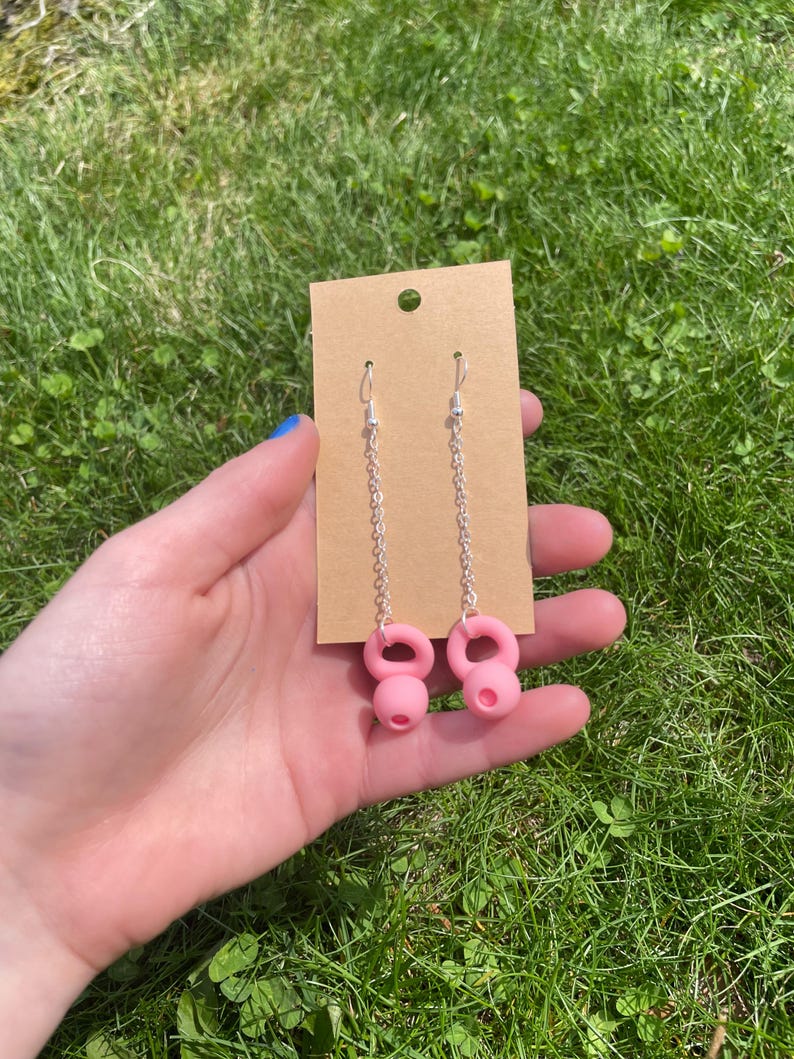 May include: A pair of dangle earrings featuring silver-toned metal chains attached to pink kettlebell-shaped charms. The earrings are displayed on a rectangular brown cardboard jewelry card with a small circular hole at the top for hanging.
