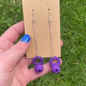 May include: A pair of drop earrings featuring purple kettlebell-shaped charms suspended from silver-toned metal chains. The earrings are attached to a rectangular tan cardboard display card with a hole at the top for hanging purposes.