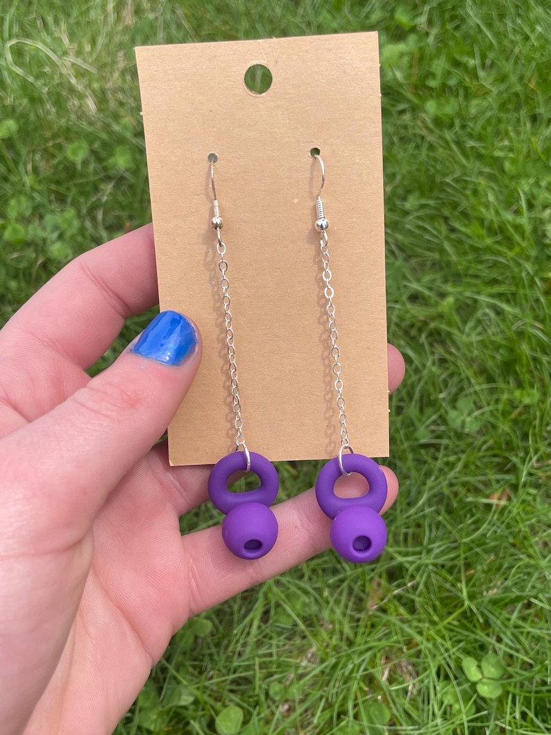 May include: A pair of drop earrings featuring purple kettlebell-shaped charms suspended from silver-toned metal chains. The earrings are attached to a rectangular tan cardboard display card with a hole at the top for hanging purposes.