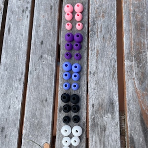 May include: A collection of silicone earbud tips arranged in a vertical column. The set includes three pairs of pink, three pairs of purple, three pairs of light blue, three pairs of black, and three pairs of white replacement ear tips for headphones.