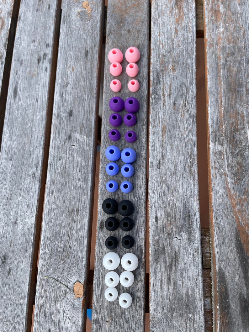 May include: A collection of silicone earbud tips arranged in a vertical column. The set includes three pairs of pink, three pairs of purple, three pairs of light blue, three pairs of black, and three pairs of white replacement ear tips for headphones.