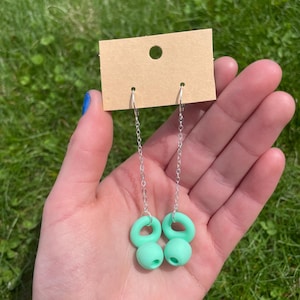 May include: A pair of dangle earrings featuring silver-toned chains attached to light mint green, rounded, donut-shaped charms. The earrings are displayed on a rectangular, tan-colored cardboard jewelry display card with a circular hole at the top.