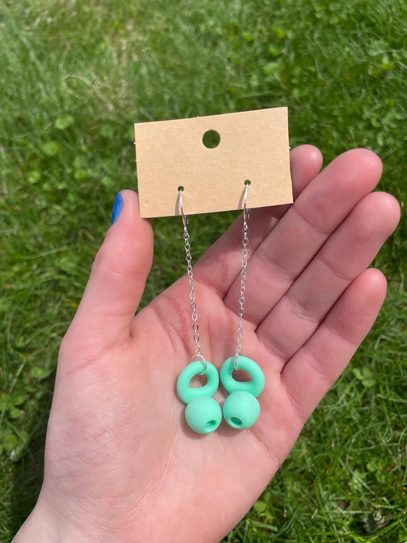 May include: A pair of dangle earrings featuring silver-toned chains attached to light mint green, rounded, donut-shaped charms. The earrings are displayed on a rectangular, tan-colored cardboard jewelry display card with a circular hole at the top.