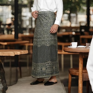 Puede incluir: Un sarong largo estampado en tonos grises y verde oliva. La tela presenta intrincados diseños geométricos y florales. El sarong se lleva con una camisa blanca y zapatos negros. El texto "TronicCraft" es visible en el sarong.