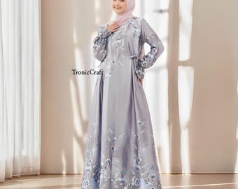 May include: A gray maxi dress with a floral print and long sleeves. The dress has a tie at the waist and a light pink headscarf.