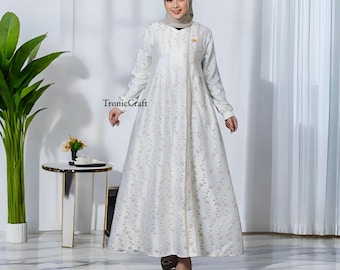 May include: A white maxi dress with a gold floral pattern and a gold trim down the front. The dress has long sleeves and a V-neckline. The model is wearing a white headscarf.