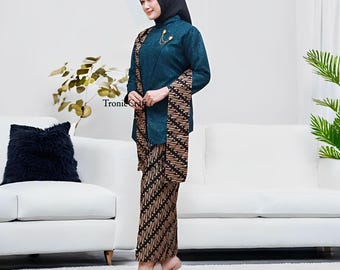 May include: A teal-colored traditional Indonesian outfit, featuring a long-sleeved top and a matching skirt with a brown batik pattern. The outfit includes a shawl with a similar batik design and a black head covering. A gold brooch accents the top.