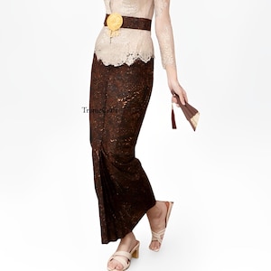 May include: A brown and gold patterned skirt with a gold belt and a white lace top. The skirt is long and flowing, and the top is fitted. The woman is wearing white shoes.