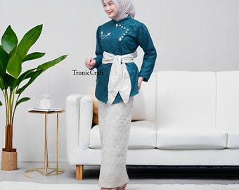 May include: Teal-colored formal outfit with a long-sleeved top and a matching skirt. The top features white floral embroidery and a white sash tied around the waist. The skirt is a light color with a floral pattern. The outfit is worn with nude heels.