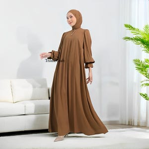 May include: A long, flowing brown dress with long sleeves and a high neckline. The dress features a button detail at the neck and ruffled cuffs. The text "Tromecraft" is visible on the dress. The model is wearing a matching head covering.
