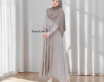 May include: A woman wearing a long, light gray dress with a tiered skirt and a matching light gray headscarf. The dress has a long sleeve and a relaxed fit. The woman is standing on a wooden floor.