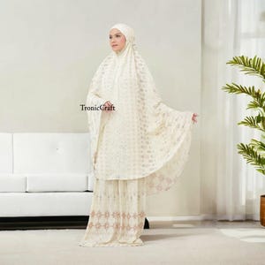 May include: A white prayer outfit with a lace pattern and a matching headscarf. The outfit has a long skirt and a loose-fitting top. The outfit is decorated with a delicate floral pattern.
