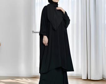 Puede incluir: Una abaya negra con una silueta larga y fluida. La abaya tiene un diseño sencillo con un cuello alto y mangas largas. La modelo lleva un pañuelo negro en la cabeza.