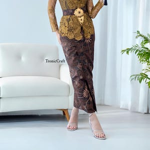 May include: A brown and gold patterned skirt with a gold belt and a gold brooch. The skirt is worn with white heeled sandals.