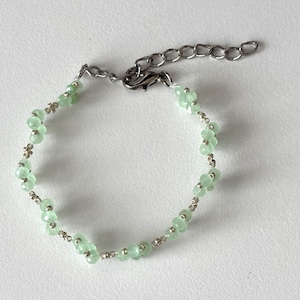 May include: A delicate bracelet featuring light green, flower-shaped beads interspersed with small silver-coloured beads. The bracelet has a silver-tone chain and clasp for adjustable sizing. Sage green, dainty, elegant, floral, hsndmade jewellery bracelet, gift for her, anniversary gift, frienship braceler, floral beaded handmade jewellery, pretty bracwlet, pastel jewellery bracelet, gifts for her, bestseller
