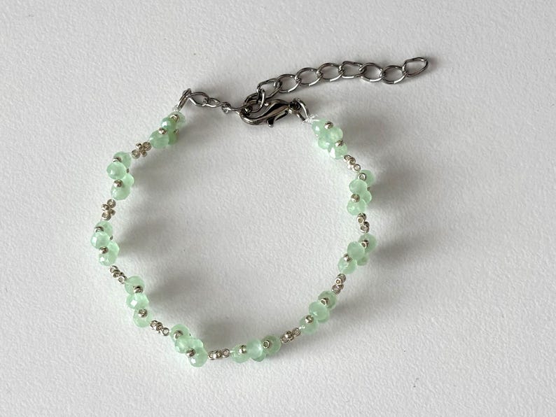 May include: A delicate bracelet featuring light green, flower-shaped beads interspersed with small silver-coloured beads. The bracelet has a silver-tone chain and clasp for adjustable sizing. Sage green, dainty, elegant, floral, hsndmade jewellery bracelet, gift for her, anniversary gift, frienship braceler, floral beaded handmade jewellery, pretty bracwlet, pastel jewellery bracelet, gifts for her, bestseller
