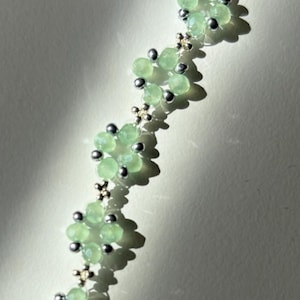 A floral bracelet with light green, faceted beads forming flower shapes. Silver-coloured beads and connectors are interspersed between the flowers. The necklace is displayed against a neutral background, highlighting the jewellery. Sage green, dainty, elegant, floral, handmade jewellery bracelet, gift for her, anniversary gift, frienship bracelet. floral beaded handmade jewellery, pretty bracelet, pastel jewellery bracelet, gifts for her, bestseller
