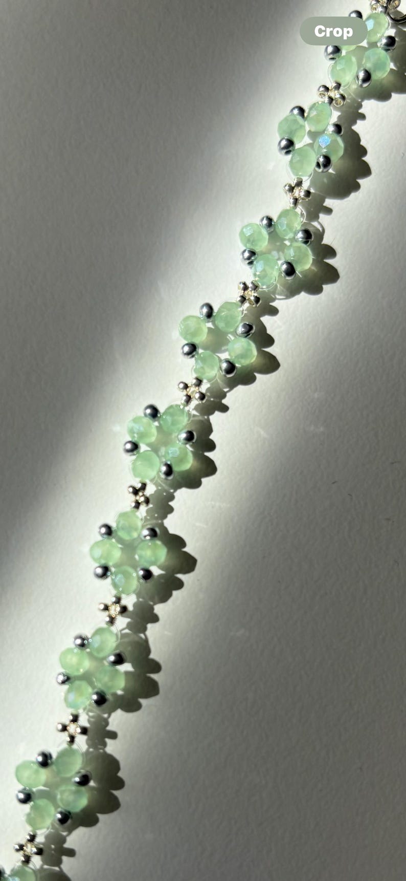 A floral bracelet with light green, faceted beads forming flower shapes. Silver-coloured beads and connectors are interspersed between the flowers. The necklace is displayed against a neutral background, highlighting the jewellery. Sage green, dainty, elegant, floral, handmade jewellery bracelet, gift for her, anniversary gift, frienship bracelet. floral beaded handmade jewellery, pretty bracelet, pastel jewellery bracelet, gifts for her, bestseller
