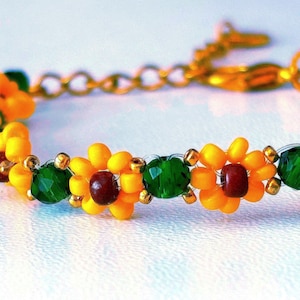 May include: A beaded bracelet featuring a repeating pattern of flower shapes constructed from yellow seed beads with dark red centers, interspersed with faceted emerald green beads and small gold-toned metallic spacer beads, finished with a gold-toned chain.