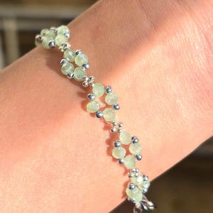 May include: A delicate bracelet featuring pale green gemstone beads and small silver-coloured accents. The bracelet is composed of multiple floral-like clusters, connected by a silver chain. Sage green, dainty, elegant, floral, handmade jewellery bracelet, gift for her, anniversary gift, frienship bracelet, floral beaded handmade jewellery, pretty bracelet, pastel jewellery bracelet, gifts for her, bestseller