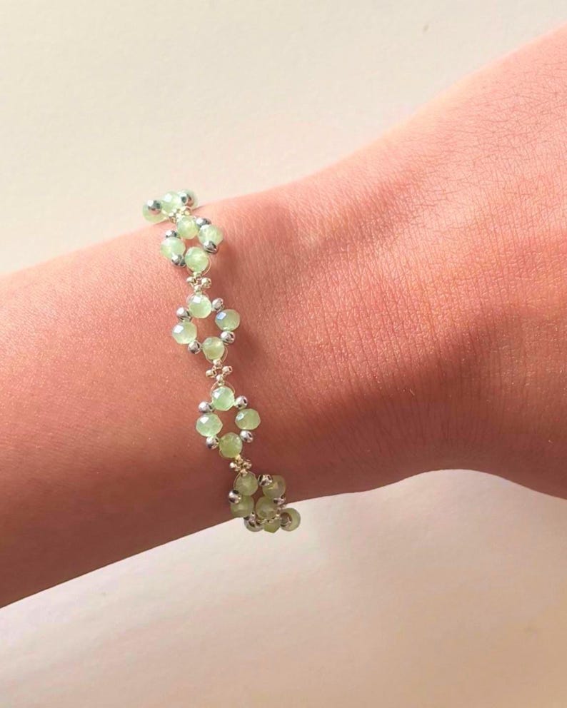 May include: A bracelet with light green beads and silver-coloured details. The beads are arranged in a repeating pattern, creating a stylish accessory. The bracelet is shown on a wrist, showcasing its design and colour. Sage green, dainty, elegant, floral, handmade jewellery bracelet, gift for her, anniversary gift, frienship braceler, floral beaded handmade jewellery, pretty bracwlet, pastel jewellery bracelet, gifts for her, bestseller