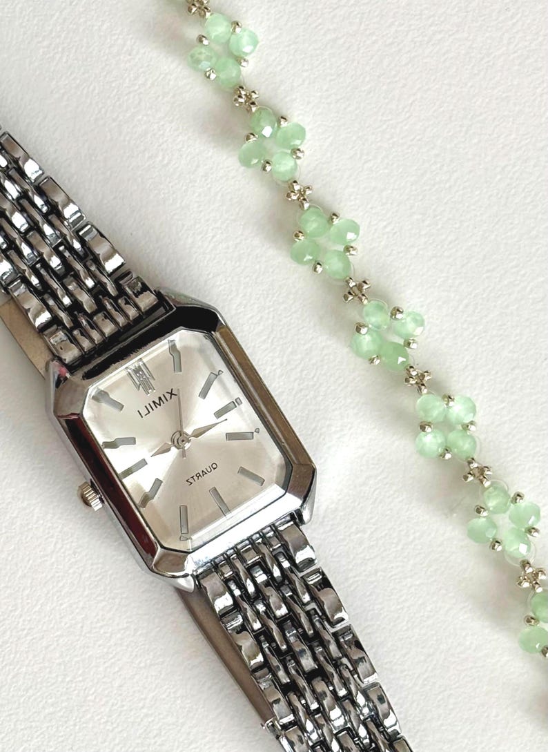 A delicate green floral bracelet is next to the watch. Sage green, dainty, elegant, floral, hsndmade jewellery bracelet, guft for her, anniversary gift, frienship braceler, floral beaded handmade jewellery, pretty bracwlet, pastel jewellery bracelet, gifts for her, bestseller