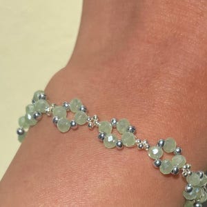May include: A bracelet with light green faceted beads and small silver beads, arranged in a floral pattern. The bracelet is worn on a wrist. The jewelry piece is made of beads and metal, and is a fashion accessory. Sage green, dainty, elegant, floral, hsndmade jewellery bracelet, gift for her, anniversary gift, frienship braceler, floral beaded handmade jewellery, pretty bracwlet, pastel jewellery bracelet, gifts for her, bestseller