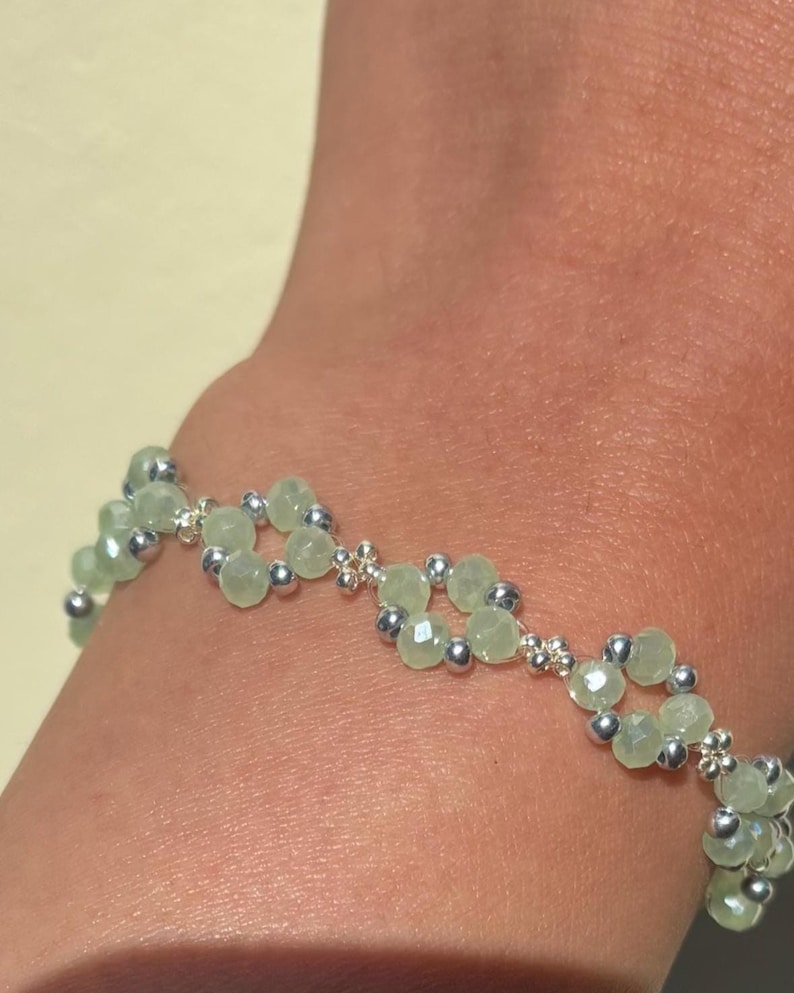 May include: A bracelet with light green faceted beads and small silver beads, arranged in a floral pattern. The bracelet is worn on a wrist. The jewelry piece is made of beads and metal, and is a fashion accessory. Sage green, dainty, elegant, floral, hsndmade jewellery bracelet, gift for her, anniversary gift, frienship braceler, floral beaded handmade jewellery, pretty bracwlet, pastel jewellery bracelet, gifts for her, bestseller