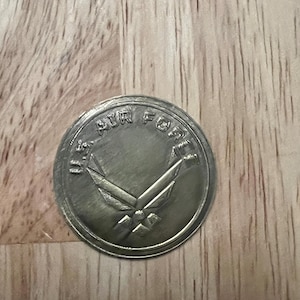 May include: A silver-colored U.S. Air Force coin. The coin features the words "U.S. AIR FORCE" around the edge and a stylized emblem in the center. The coin is circular and has a slightly worn appearance.