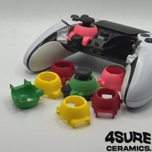 May include: A white video game controller with a pink joystick ring installed, accompanied by several loose, colorful plastic joystick rings in red, yellow, and green. The text 4SURE CERAMICS is printed in black in the bottom right corner.
