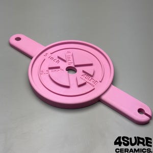May include: A light pink circular ceramic centering tool with two extended handles featuring holes at each end. The surface displays embossed text reading 3.5 inch, 4 inch, 5 inch, and the brand name ASURE CERAMICS with a central circular opening.