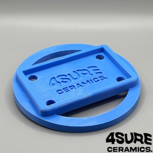 May include: A bright blue 3D printed object featuring a circular base with a rectangular frame attached to the top. The rectangular frame contains four circular holes and the embossed text 4SURE CERAMICS. The surface has a visible layered texture.