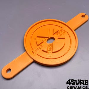 Op de afbeelding: Een oranje, cirkelvormig keramisch centreergereedschap met twee verlengde handvatten met gaten aan de uiteinden. Het oppervlak toont in reliëf gedrukte tekst 4 inch, 5 inch, 3.5 inch, de merknaam 4SURE CERAMICS en een centraal gat.