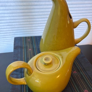 May include: A yellow-green ceramic teapot with a matching lid and a curved handle sits in front of a matching pitcher. The pitcher has a tall, slender shape with a curved spout and a handle. Both pieces are on a patterned surface.