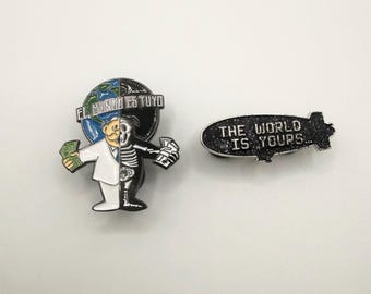 May include: Two enamel pins. One pin features a split design of a man holding money, half human and half skeleton, with the text "EL MUNDO ES TUYO" above. The other pin is a black blimp with the text "THE WORLD IS YOURS..."