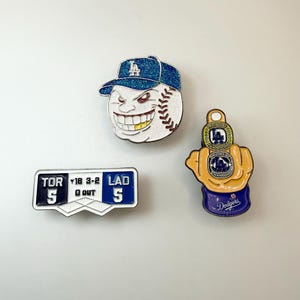 May include: Three baseball-themed enamel pins. One pin features a baseball with a blue cap and a grinning face. Another pin is a scoreboard with "TOR" and "LAD" on it. The third pin is a baseball glove with a ring that says "World Champions".