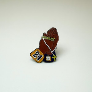 May include: A brown pin featuring clasped hands in prayer, adorned with a beaded bracelet and a rosary. The pin includes a gold and blue emblem with the number 24, and another with the number 8 and a cross.