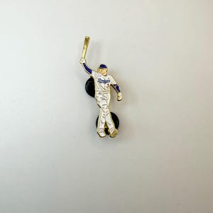 Pu&ograve; includere: Spilla da baseball raffigurante un giocatore in uniforme bianca con dettagli blu e rossi, che tiene una mazza d'oro. La spilla presenta la scritta "Dodgers" ed &egrave; attaccata a un supporto nero.