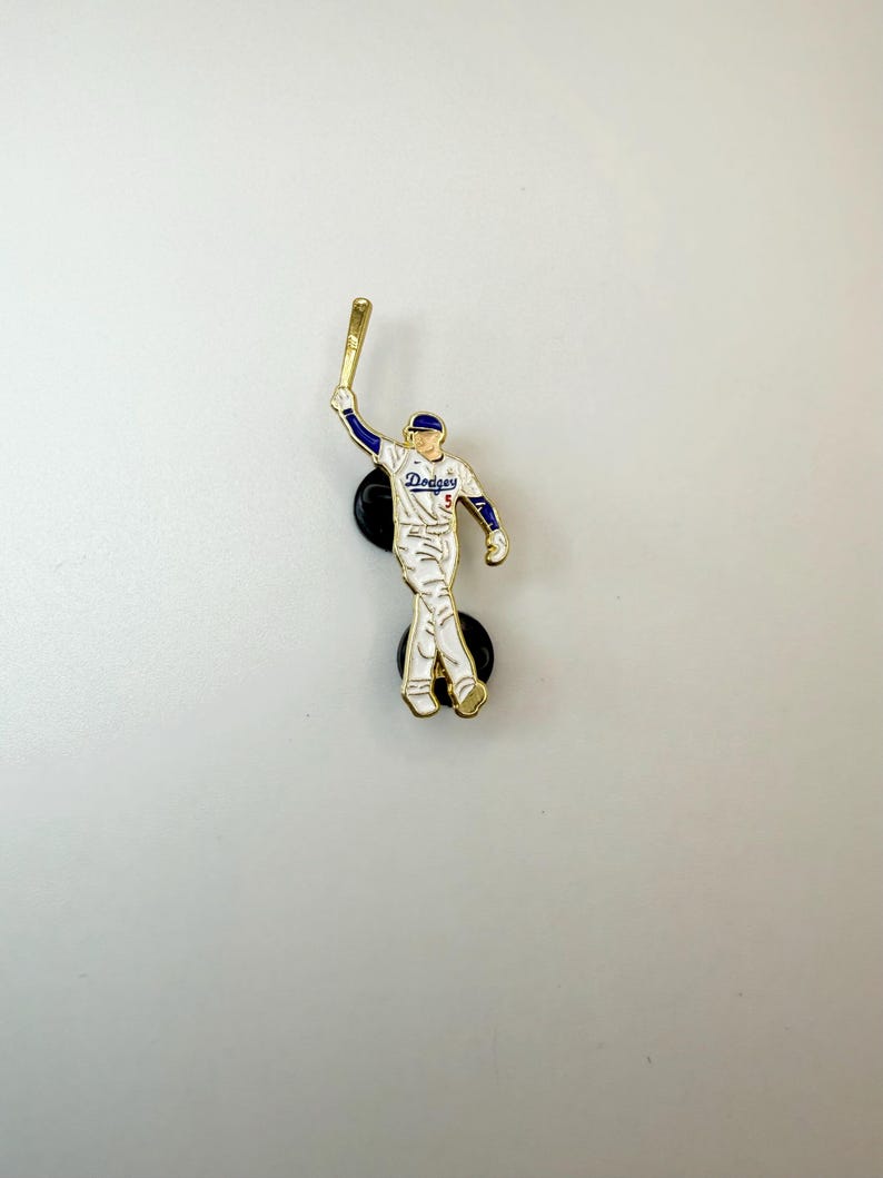Pu&ograve; includere: Spilla da baseball raffigurante un giocatore in uniforme bianca con dettagli blu e rossi, che tiene una mazza d'oro. La spilla presenta la scritta "Dodgers" ed &egrave; attaccata a un supporto nero.