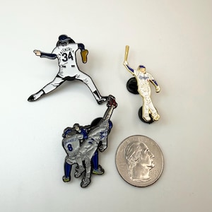 Pu&ograve; includere: Tre spille smaltate a tema baseball. Una spilla raffigura un lanciatore in uniforme bianca con il numero 34. Un'altra mostra un battitore in uniforme bianca e blu. La terza spilla mostra due giocatori in uniforme blu. Una moneta &egrave; inclusa per la scala.