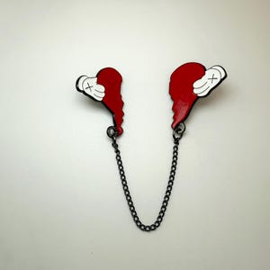 May include: A pair of red heart-shaped pins connected by a black chain. Each pin features a white glove with black details. The pins are on a white background.