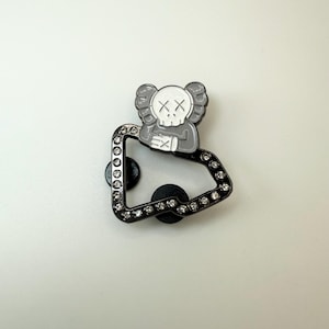 May include: A black and silver pin featuring a stylized skull character with crossed-out eyes and a black square frame adorned with small, clear crystals. The pin has two black pin backs.