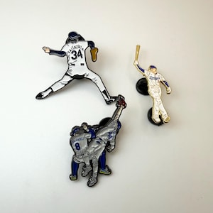 Pu&ograve; includere: Tre spille smaltate a tema baseball. Una spilla raffigura un giocatore in posa da lancio, un'altra un battitore e la terza due giocatori che festeggiano. Le spille sono bianche, blu e oro.