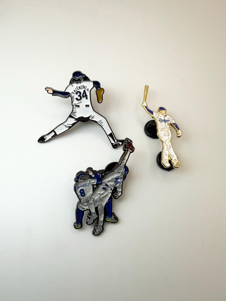 Pu&ograve; includere: Tre spille smaltate a tema baseball. Una spilla raffigura un giocatore in posa da lancio, un'altra un battitore e la terza due giocatori che festeggiano. Le spille sono bianche, blu e oro.