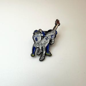 Pu&ograve; includere: Spilla a tema baseball con due giocatori in azione. Un giocatore si allunga per prendere una palla. La spilla ha una combinazione di colori argento e blu, con il numero 8 visibile sulla divisa di un giocatore. Circa 3 cm di altezza.