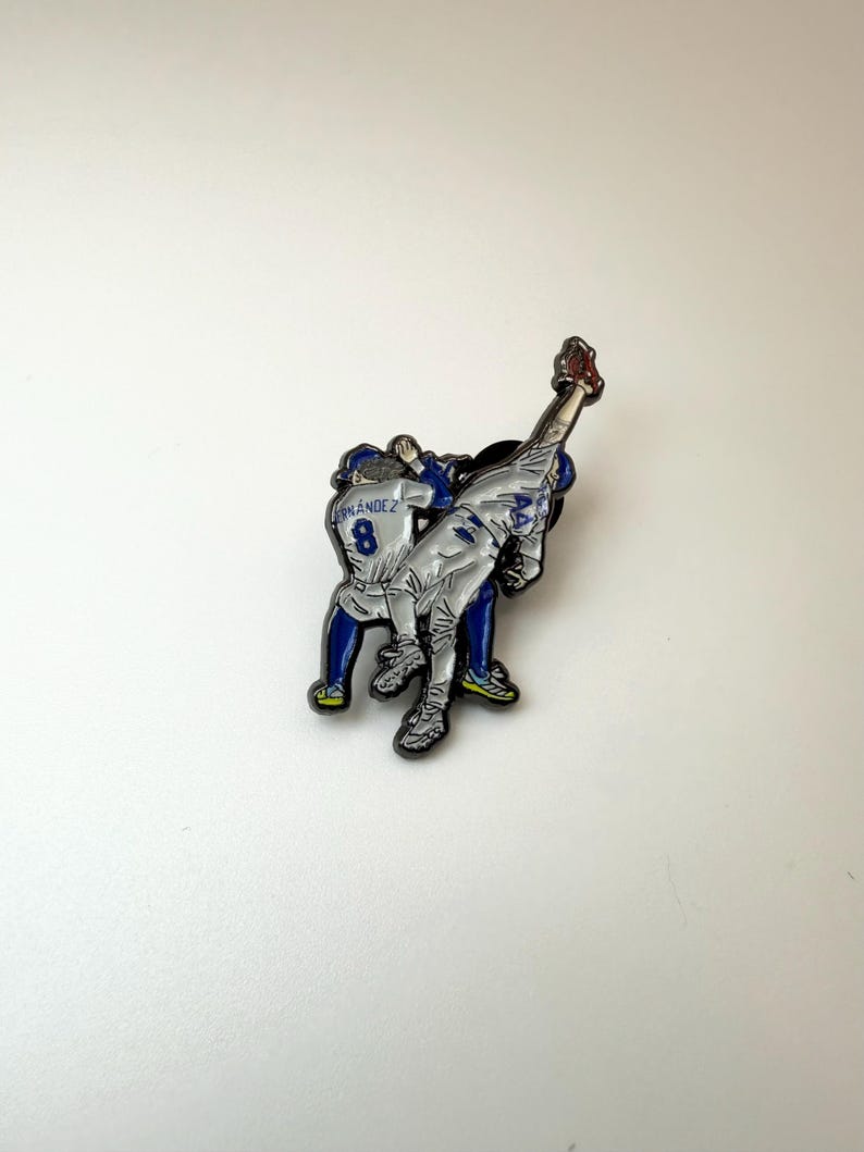 Pu&ograve; includere: Spilla a tema baseball con due giocatori in azione. Un giocatore si allunga per prendere una palla. La spilla ha una combinazione di colori argento e blu, con il numero 8 visibile sulla divisa di un giocatore. Circa 3 cm di altezza.